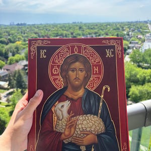 May include: A religious icon depicting Jesus holding a lamb, with a red border and gold detailing. Jesus is shown in a red robe and blue cloak. The background shows a city landscape. The icon has gold lettering.