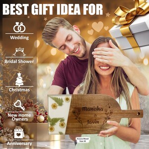 May include: A personalized acacia wood serving board with the names "Manisha & Javin" engraved on it. The board is decorated with pressed flowers and has a light green and white resin inlay. The board is a great gift idea for a wedding, bridal shower, anniversary, or new home.