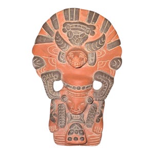 Op de afbeelding: Een terracotta-gekleurde, gebeeldhouwde sculptuur van een gestileerde figuur. De sculptuur toont een centraal figuur met een lachend gezicht, omringd door ingewikkelde ontwerpen en patronen. Het stuk heeft een platte achterkant en is waarschijnlijk een wandkleed.
