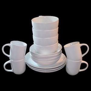 Puede incluir: Un juego de vajilla blanco, que incluye cuencos apilados, platos y tazas con asas curvas. El conjunto está dispuesto sobre un fondo negro, mostrando una estética limpia y minimalista. La vajilla es de cerámica.