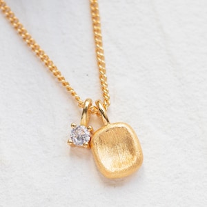 May include: A gold chain necklace with a small, square pendant. The pendant has a brushed finish and a small, clear rhinestone.