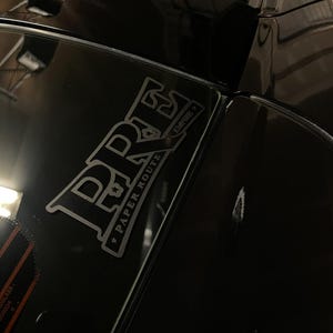 Può includere: Un'auto nera con un adesivo argentato sul lunotto posteriore. L'adesivo dice "PRS Paper Route Empire".