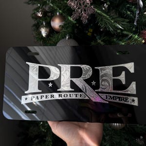 Puede incluir: Placa de matrícula negra con un diseño metálico plateado. El diseño presenta las letras "PRE" con las palabras "PAPER ROUTE EMPIRE" debajo.