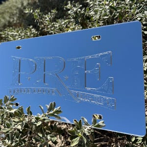 May include: A silver metal license plate with the word "PRE" in a decorative font. The plate has two holes on the top and two holes on the bottom.