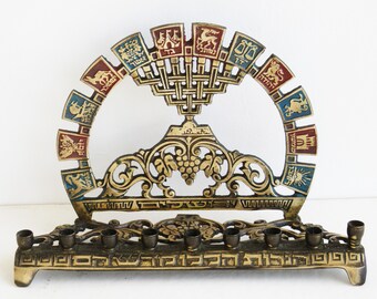 May include: A gold menorah with a decorative design featuring a grapevine and Hebrew text. The menorah has nine candle holders and a central shamash holder. The menorah is decorated with colourful enamel and gold accents.