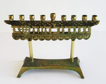 May include: A vintage brass menorah with nine candle holders. The menorah features a decorative design with Hebrew lettering and is supported by two brass legs and a base. The base has the word "Jerusalem" engraved on it.
