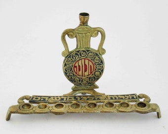 May include: A brass menorah with a decorative, round, gold-coloured vase with a red and black design. The vase has a circle with Hebrew text and symbols around it. The menorah has eight candle holders and Hebrew text on the base.