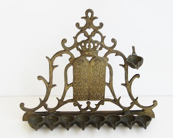 May include: A brass menorah with a decorative design featuring a crown and the Ten Commandments. The menorah has nine candle holders and a single oil lamp on the side.