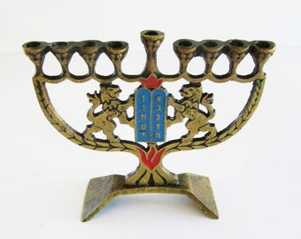 May include: A brass-coloured menorah with seven candle holders. The base has a rectangular shape. The menorah is decorated with two lions, a blue tablet with Hebrew text, and red and gold accents.