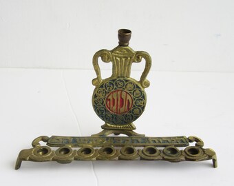 May include: A brass menorah, a Jewish candelabrum, with a central vase-shaped element and a base with nine candle holders. The vase has a blue and red circular design with Hebrew text. Used during the Hanukkah festival.