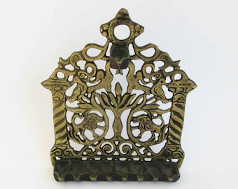 May include: A brass Hanukkah menorah featuring an intricate openwork design with two lions flanking a central pillar, topped by a circular hanging loop. The base includes eight small, aligned oil cups for lighting, all crafted in a metallic gold tone.