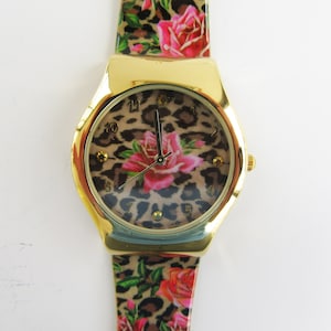 Può includere: Un orologio da polso dorato con un design a stampa leopardata e rosa rosa sul quadrante e sul cinturino.
