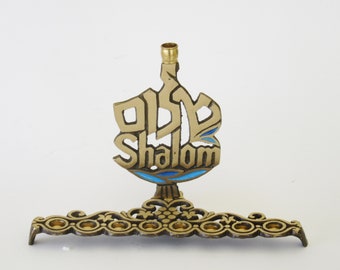 May include: A gold menorah with a Shalom inscription in blue enamel. The menorah has a decorative base with a grapevine design.