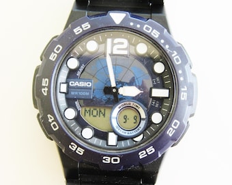 Könnte beinhalten: Eine schwarze Casio Armbanduhr mit einem blauen Zifferblatt und einer Weltzeitfunktion. Die Uhr verfügt über ein digitales Display, das den Wochentag und die Uhrzeit anzeigt. Die Uhr hat außerdem eine 10-jährige Batterie.