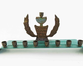 May include: A vintage menorah with a green metal base and a gold-toned menorah with a wreath design. The menorah has nine candle holders.