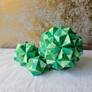 Pode incluir: Três esculturas geométricas de origami de tamanhos variados, construídas com papel dobrado em vários tons de verde, variando do verde-limão claro ao verde-floresta profundo, com facetas triangulares e pentagonais intrincadas e interligadas.