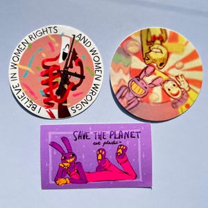Op de afbeelding: Drie stickers: een ronde met een rood lint, een geweer en de tekst I BELIEVE IN WOMEN RIGHTS AND WOMEN WRONGS; een ronde met stripfiguren op een zonnestraal-achtergrond; en een rechthoekige paarse met een konijn en SAVE THE PLANET eat plastic.