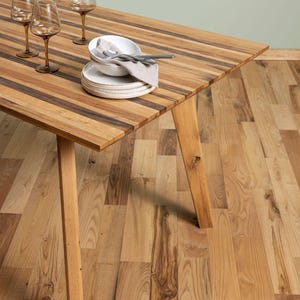 Könnte beinhalten: Ein Esstisch aus Holz mit einem gestreiften Tischplattendesign, das abwechselnd helle und dunkle Holztöne aufweist. Der Tisch ist mit weißen Tellern, Besteck, einer Leinen-Serviette und drei Weingläsern gedeckt. Die Tischbeine sind abgewinkelt und aus hellem Holz gefertigt.