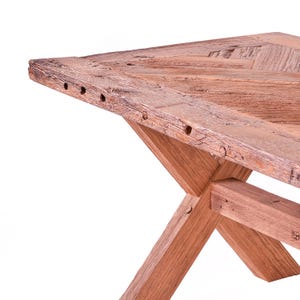 Op de afbeelding: Close-up van een rustieke houten tafel met een verweerde afwerking. De tafel heeft een kruisvormige basis en een verweerd tafelblad met zichtbare houtnerf en spijkergaten. De natuurlijke houttinten creëren een warme, uitnodigende esthetiek.