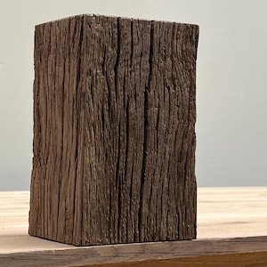 May include: A weathered, rectangular wooden block with a rough, textured surface. The block is a dark brown color, with visible grain and vertical lines. The wood appears aged and rustic, suggesting a natural or reclaimed material.