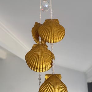May include: A decorative mobile with four gold scallop shells hanging from a wooden ring and crystal. The shells are strung with white pearls. The shells are painted gold and are suspended in a vertical arrangement.