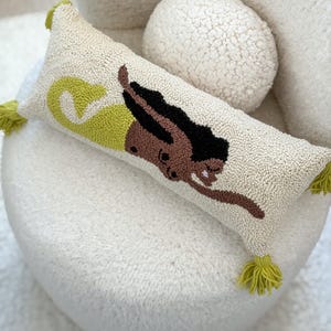 May include: Cream-colored rectangular pillow with a tufted design featuring a woman swimming. The woman has brown skin, black hair, and is wearing a yellow swimsuit. The pillow has green tassels on each end.