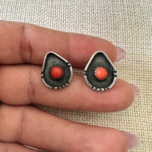 May include: A pair of silver earrings with a teardrop shape and a small red coral stone in the center.