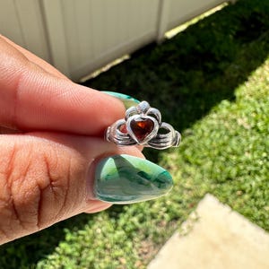 Peut inclure: Une bague en argent avec une pierre rouge en forme de cœur au centre. La bague est conçue avec un dessus en forme de couronne et une bande de nœud celtique.