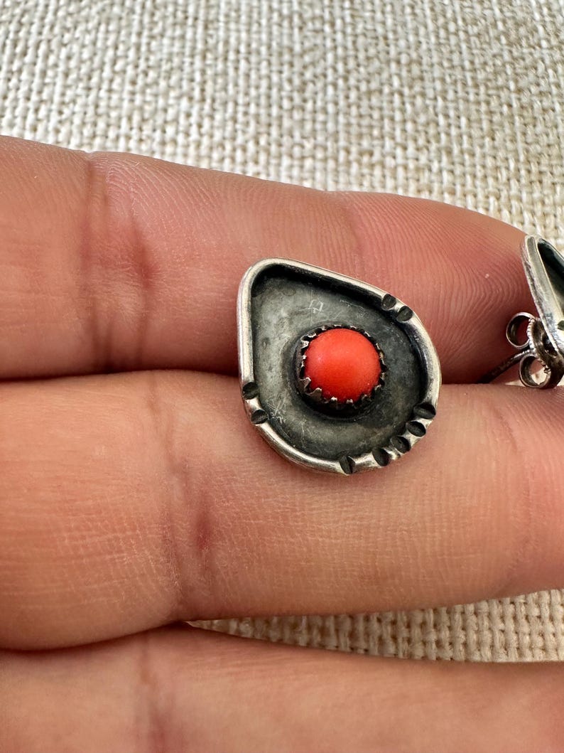 May include: A pair of silver earrings with a coral stone in the center. The earrings are in the shape of a teardrop and have a textured design.