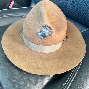 Puede incluir: Un sombrero de fieltro marrón con ala ancha y una cinta beige. El sombrero presenta un emblema metálico en la parte delantera. El sombrero está sobre una superficie de cuero negro.