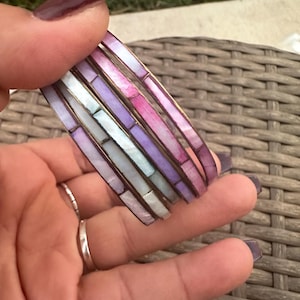 May include: A set of six iridescent bangles with a mosaic pattern of purple, pink, and white. The bangles are made of mother of pearl and have a silver finish.