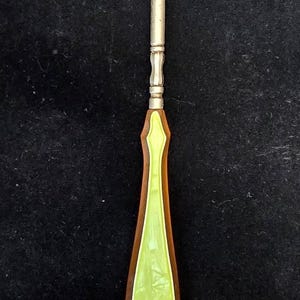 May include: A button hook featuring a metallic silver-toned shaft with a curved hook at the top and a tapered handle. The handle is constructed with a dark brown outer frame and a smooth, light lime green inlay that runs down the center.
