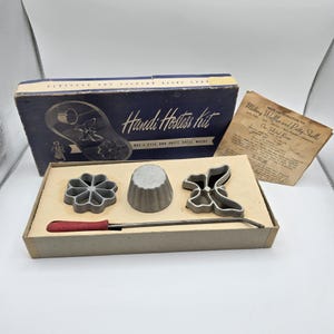 May include: A Handi Hostess kit featuring a blue box with white text, a recipe instruction sheet, and metal molds including a flower shape, a cup shape, and a butterfly shape. A long metal handle with a red textured grip is included for the molds.