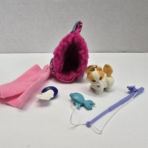 Puede incluir: Colección de juguetes en miniatura, que incluye una bota rosa con puño estampado, una bufanda rosa, una figura de gato blanco y marrón, un pez azul, un objeto azul y blanco y una caña de pescar morada.