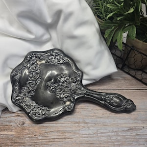 May include: A silver-toned hand mirror featuring an ornate, raised floral relief pattern across the back and handle. The metal surface displays a dark, aged patina with intricate sculpted flower and leaf details surrounding the central area.