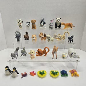 Puede incluir: Una colección de figuras de animales en miniatura, incluyendo perros, gatos, un panda, un conejo y un oso, exhibidas en estantes transparentes escalonados. Las figuras están pintadas en varios colores y estilos, mostrando una variedad de animales.