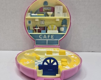 May include: A vintage pink and yellow Polly Pocket cafe playset, open to reveal a miniature interior with a cafe sign, furniture, and a blue door. The playset is in the shape of a rounded shell.