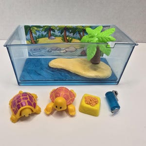 May include: A clear plastic terrarium with a tropical island scene, including palm trees and blue water. Two toy turtles, a yellow food block, and a blue water bottle are in front of the terrarium. The turtles are yellow and orange.