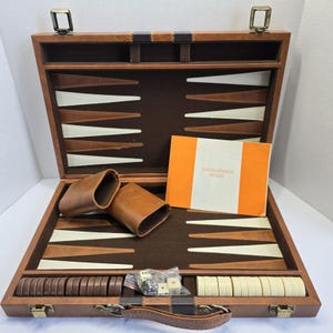 Puede incluir: Juego de backgammon abierto en un estuche de cuero marrón. El juego incluye fichas marrones y blancas, dados y un libro de reglas. El interior está forrado con fieltro marrón. Medidas: aprox. 40 cm.