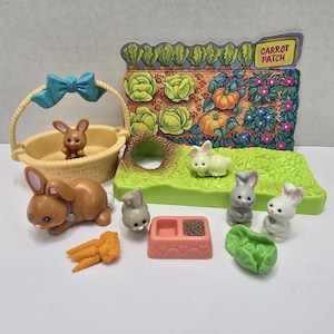 Puede incluir: Colección de conejitos de juguete en miniatura, una cesta con lazo azul, zanahorias, un cuenco para comida y una escena de jardín con la inscripción "Carrot Patch". Los conejitos son de varios colores, incluyendo marrón, gris y blanco.