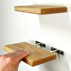 May include: Two rectangular light brown wooden shelves with a visible grain pattern. Each shelf is mounted to the wall using two silver-colored metal brackets that feature rectangular base plates secured with screws at each end of the metal support.