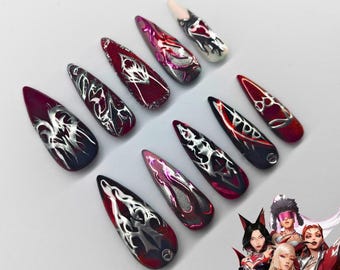 May include: A set of ten stiletto-shaped press-on nails featuring a deep red and black base with metallic silver liquid-style designs. The nails display intricate, flowing chrome patterns and glossy finishes. A small illustration of four anime-style characters in red and silver outfits is positioned in the bottom right corner.