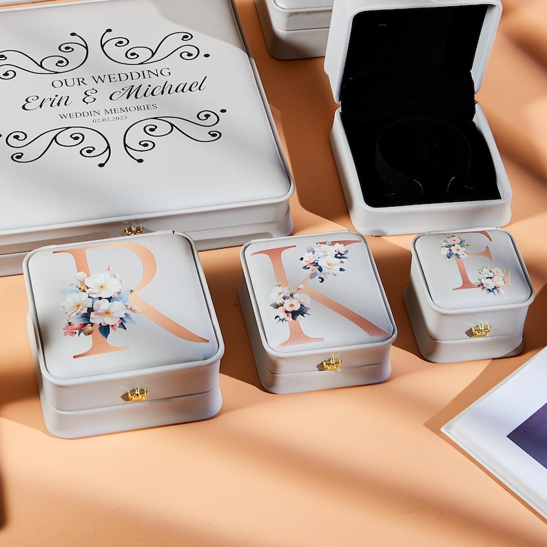 May include: A collection of gray jewelry boxes with floral and initial designs. The boxes feature gold-colored clasps and are arranged on a peach-colored surface. One box is open, revealing a black interior.
