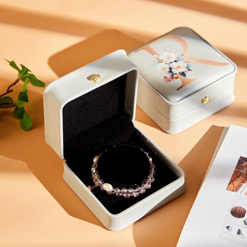 May include: A white jewelry box with a black interior holds a beaded bracelet with pearls and purple beads. The box is open, revealing the bracelet. The lid of the box features a floral design and the letter "R".