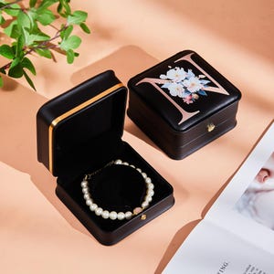 Może przedstawiać: Dwa czarne pudełka na biżuterię. Jedno otwarte, ukazujące perłową bransoletkę na czarnej aksamitnej poduszce. Drugie zamknięte, z kwiatowym wzorem i literą "N". Oba mają złote obramowania. Na różowej powierzchni.