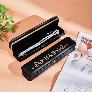 Peut inclure: Deux boîtes à bijoux noires, dont une ouverte révélant un collier de perles blanches. L'autre boîte fermée porte les initiales "H&B" en or rose. Les boîtes sont posées sur un fond pêche.