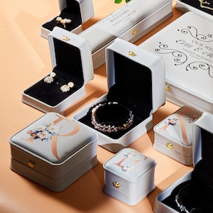 May include: An assortment of white jewelry boxes, some open to reveal earrings and bracelets. The boxes feature floral designs, initials, and gold-colored clasps. Some boxes have text that reads "Williams Always & Forever" and "Our Wedding Memories".