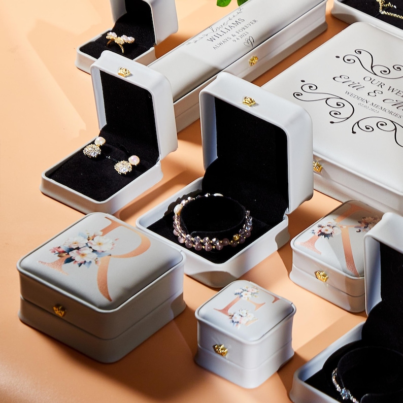 May include: An assortment of white jewelry boxes, some open to reveal earrings and bracelets. The boxes feature floral designs, initials, and gold-colored clasps. Some boxes have text that reads "Williams Always & Forever" and "Our Wedding Memories".