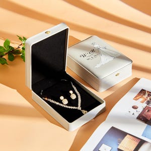 May include: A white jewelry box with a black interior and a silver clasp. The box is open and contains a silver necklace and earrings with clear stones. The box has a white lid with a bride and groom illustration and the text "W-M Mr & Mrs 15.10.2021".