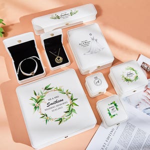 May include: An assortment of white jewelry boxes with gold clasps, some open to reveal jewelry. The boxes feature floral designs, wedding details, and the names "Smithson" and "Mr & Mrs". The boxes are arranged on a peach-colored surface.